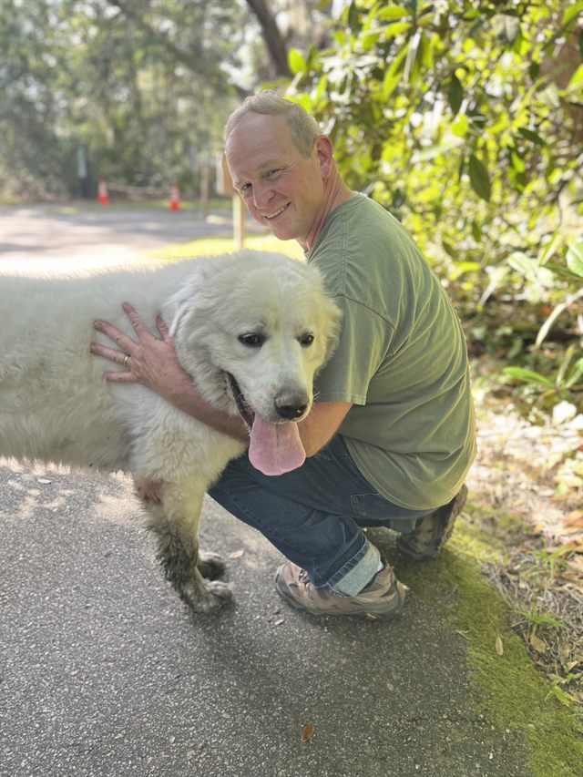 Joseph Styons kneeling on a shaded road beside a large white dog.